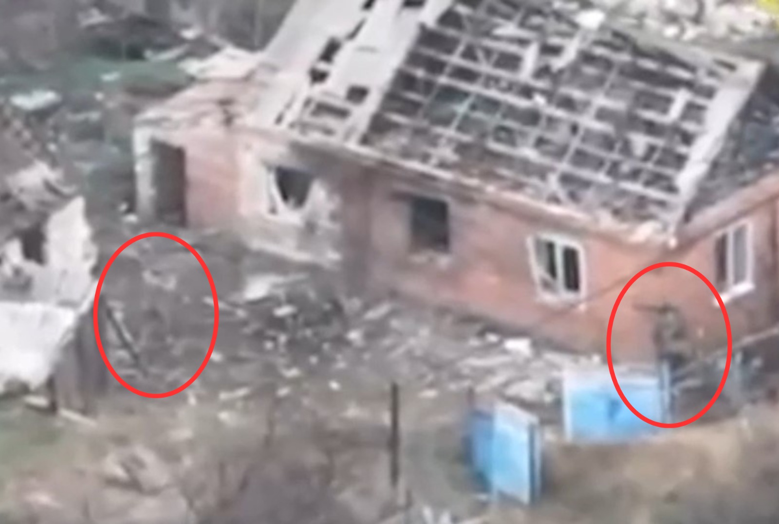 Aerial surveillance footage shows two figures marked with red circles standing amid destroyed buildings with collapsed roofs and blown-out windows, with debris scattered throughout the devastated urban area. The grainy image captures extensive structural damage to multi-story residential buildings, including a prominent pink-walled structure missing its roof.
