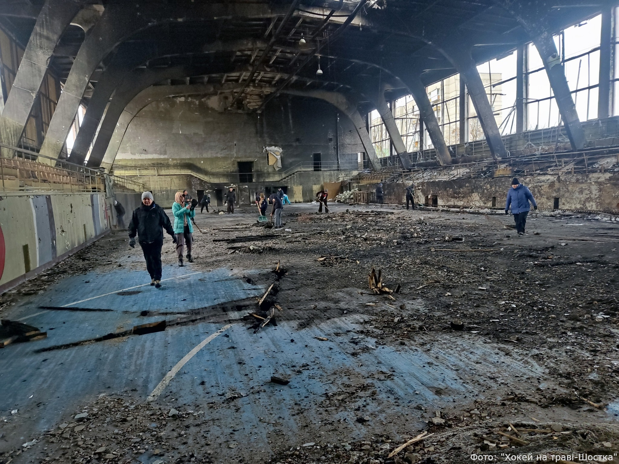 People walk through a severely damaged large industrial or public building with exposed arched ceiling structures, broken windows, and debris-covered floors. The space shows extensive destruction with collapsed roof sections and scattered rubble throughout the interior. Photo credit: Hockey on the Grass - Shostka.