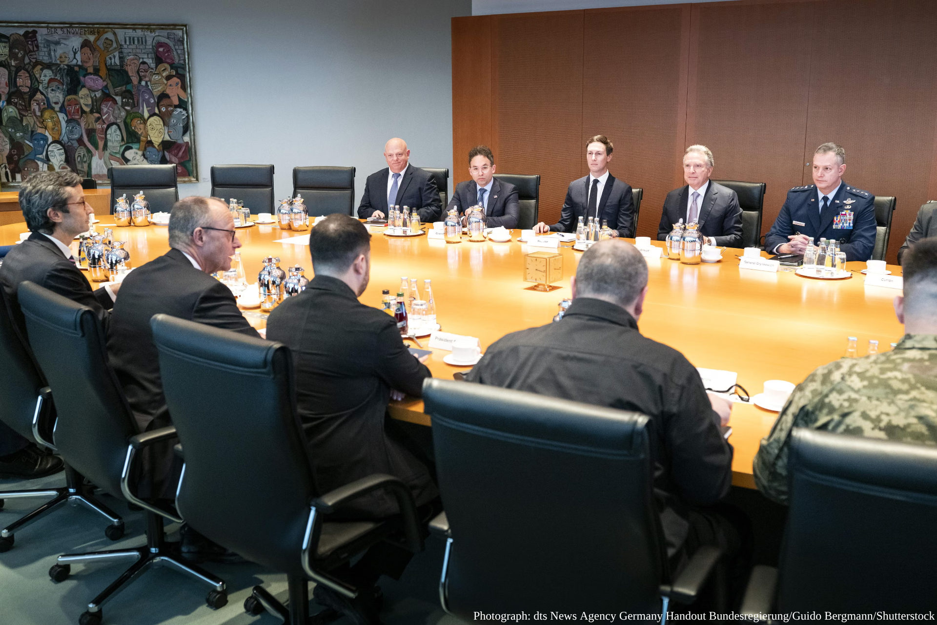 Government and military officials in formal attire sit around a polished wooden conference table with water bottles and documents during a meeting in Adlon Hotel in Berlin, with a colorful artwork visible on the wall behind them, photographed by dts News Agency Germany for Bundesregierung.
