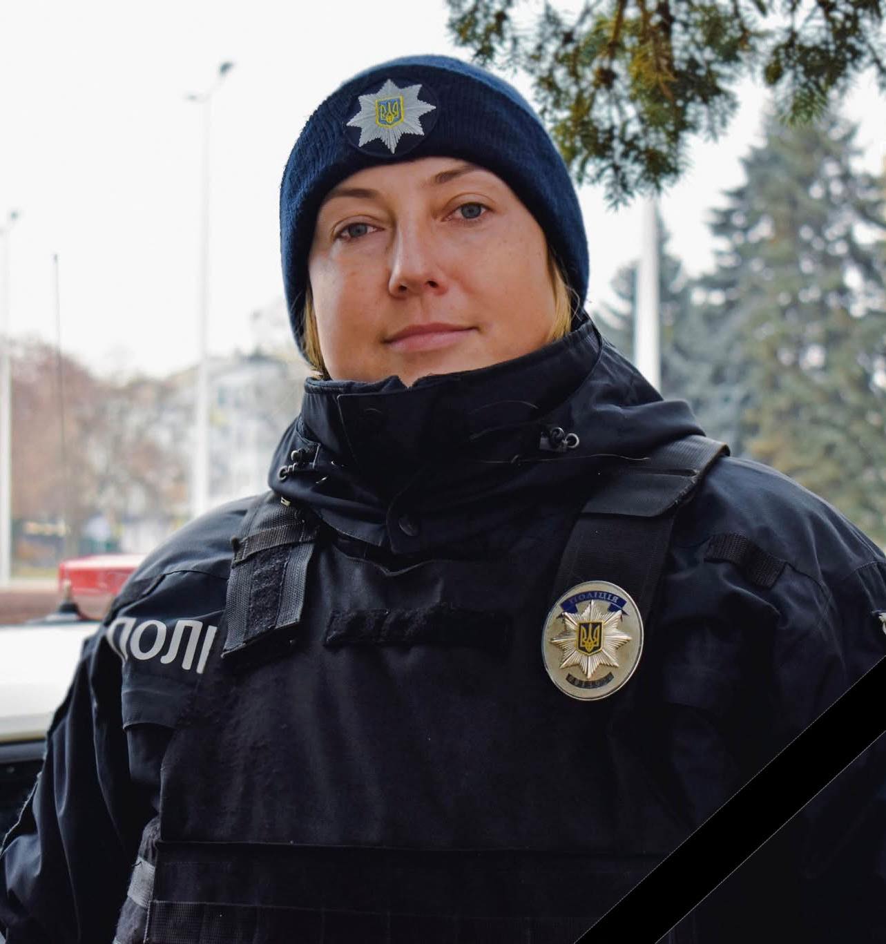 A Ukrainian police officer in tactical uniform stands outdoors in winter, wearing a dark blue knit cap with a police star badge and a black tactical vest displaying a circular police emblem. Behind her, evergreen trees and urban buildings are visible in the overcast daylight.