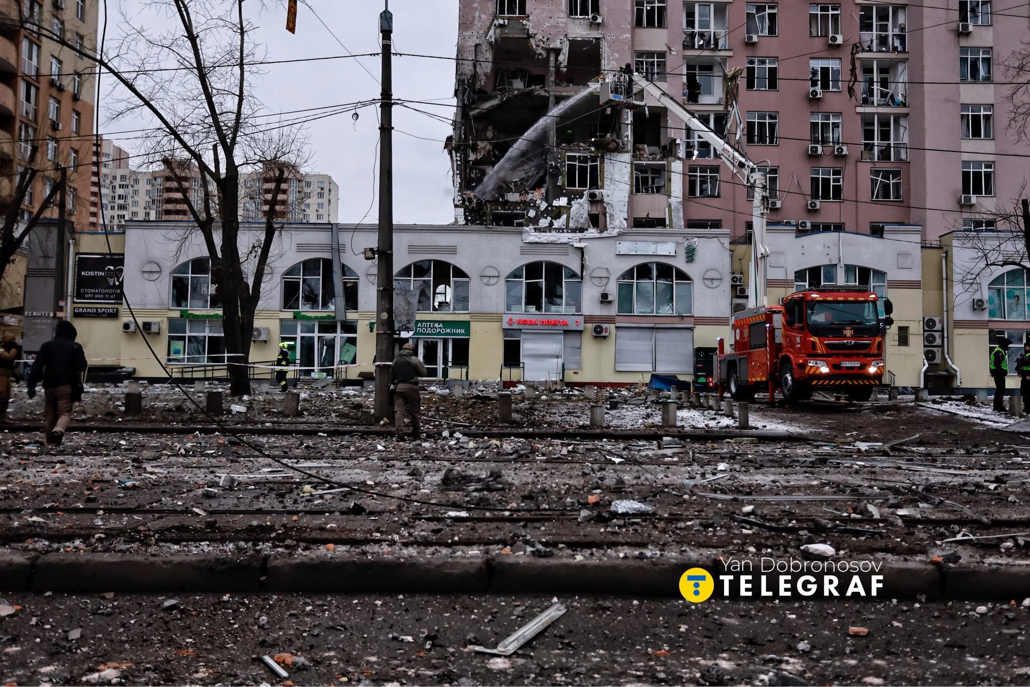 A severely damaged multi-story building with its upper floors completely destroyed stands above ground-level storefronts, while a red fire truck with an extended ladder works at the scene and emergency personnel in reflective vests move through debris-strewn streets. The devastated urban area shows extensive structural damage to residential buildings, scattered rubble across the ground, and bare winter trees amid the destruction.