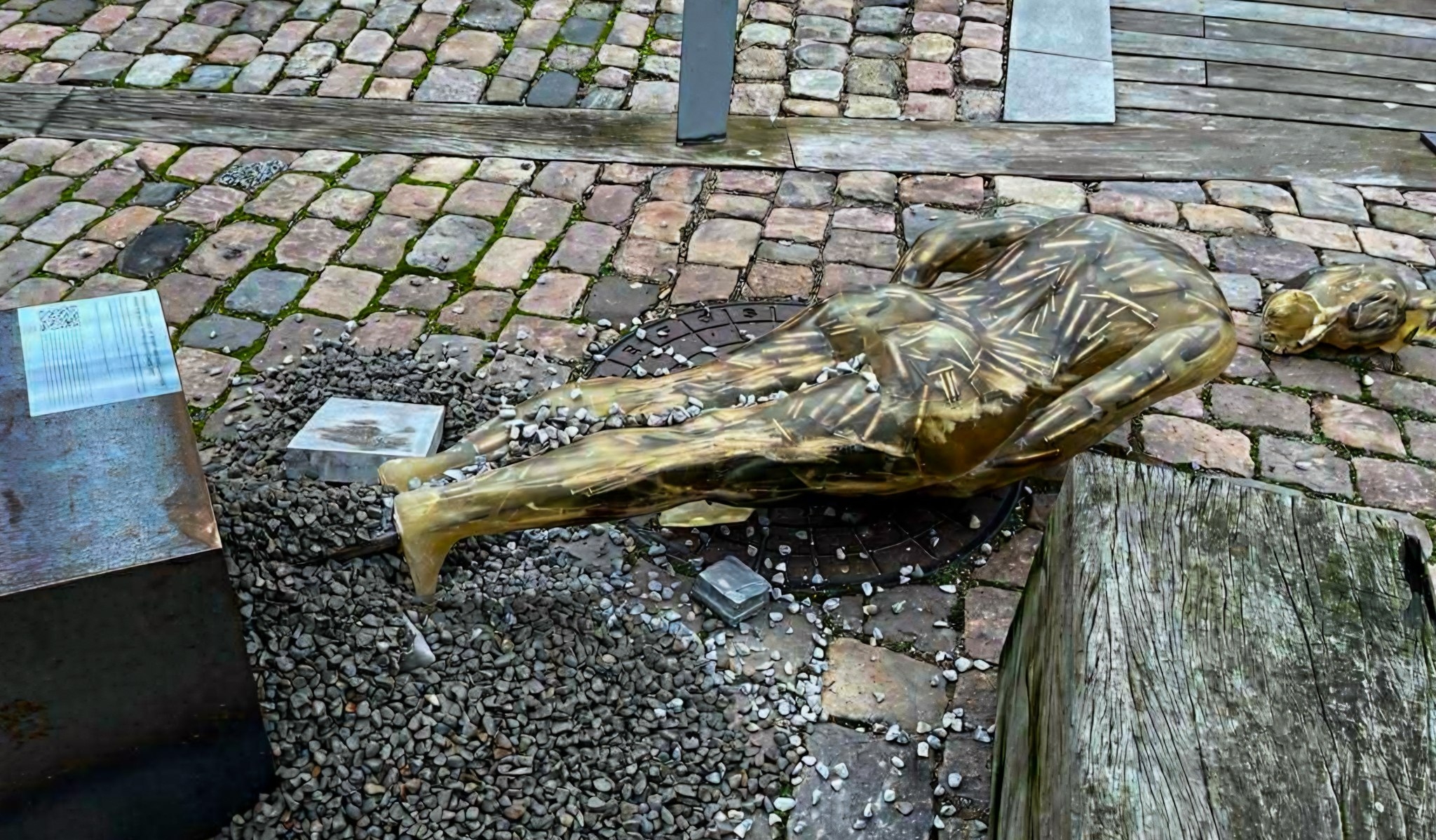 A bronze memorial sculpture of a person lying face-down rests on a bed of dark stones set into cobblestone pavement, with informational plaques visible nearby and wooden planks in the foreground. The fallen figure memorial is surrounded by scattered white stones or pebbles as a form of remembrance tribute.