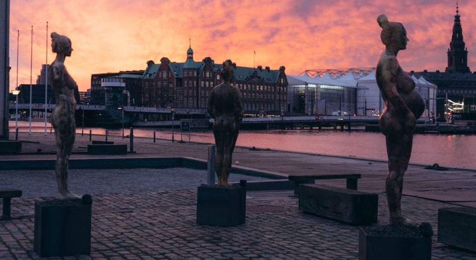 Three bronze statues stand on pedestals along a waterfront promenade at sunset, with historic buildings featuring traditional Danish architecture visible across the harbor under a pink and purple twilight sky. The silhouetted figures face the water, creating a contemplative scene against Copenhagen's illuminated cityscape.