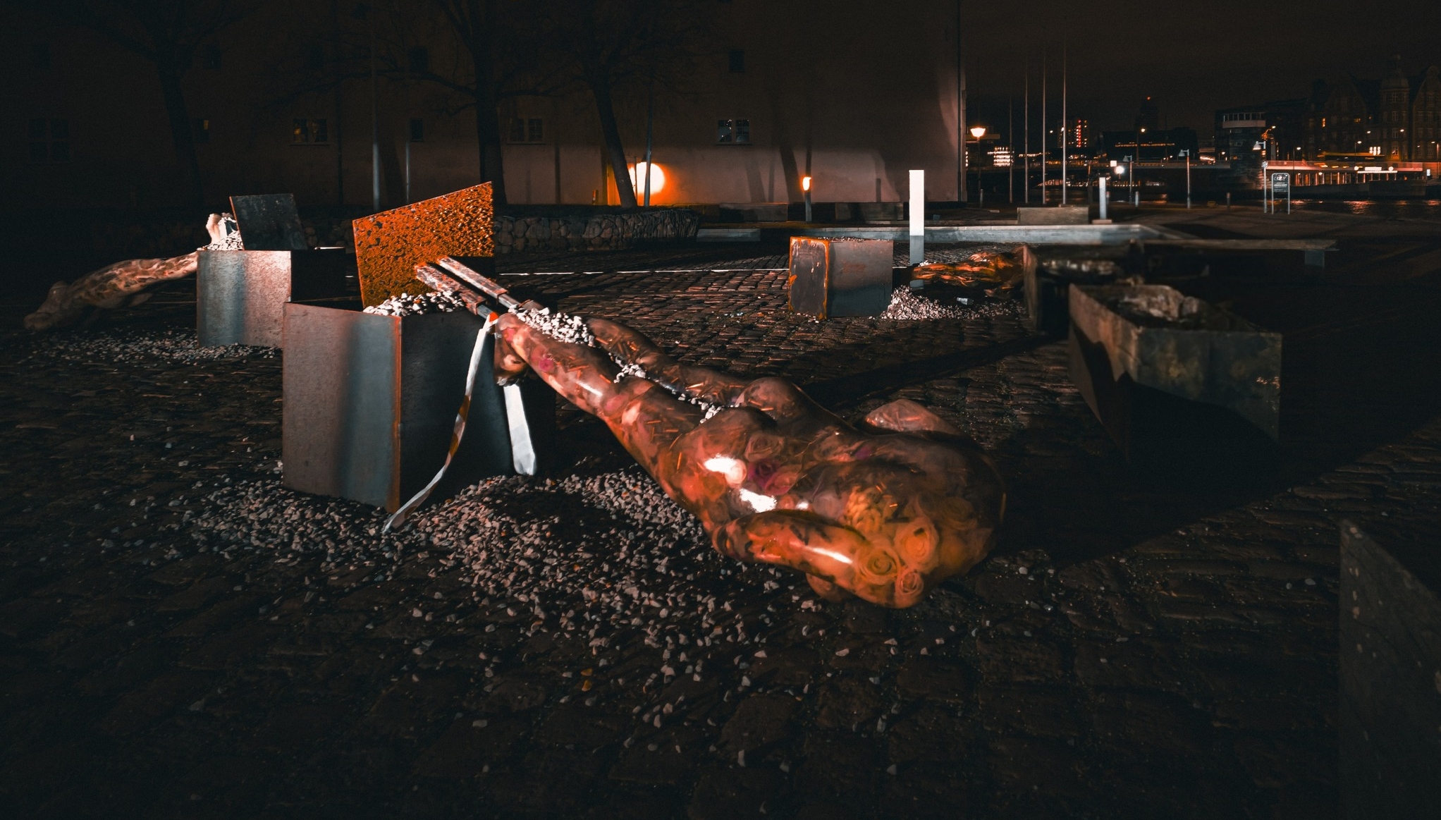 A nighttime scene shows an illuminated art installation of a large sculptural hand with glowing elements resting on dark gravel, accompanied by damaged benches and metal grating, with street lights and buildings visible in the background. The weathered copper or bronze hand sculpture appears to be part of a memorial or public art piece in an urban waterfront setting.