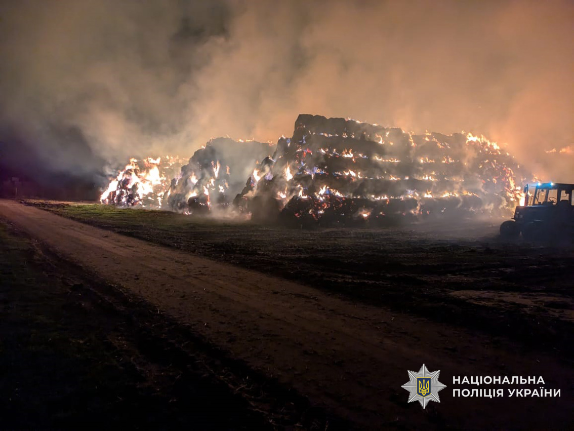 A massive burning haystack illuminates the night with orange flames and thick smoke billowing into the dark sky due to Russian attack on Ukraine, while a tractor is silhouetted on the right side of the dirt road, documented by the National Police of Ukraine.