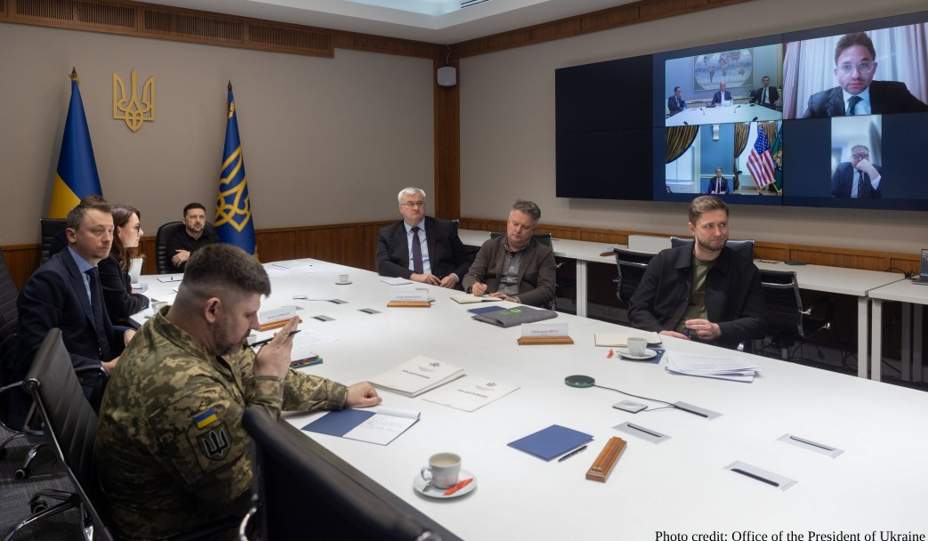 Government and military officials gather around a white conference table in a formal meeting room, with the Ukrainian flag and coat of arms displayed on the wall. A large screen shows remote participants joining via video conference, including officials with the American flag visible in the background.