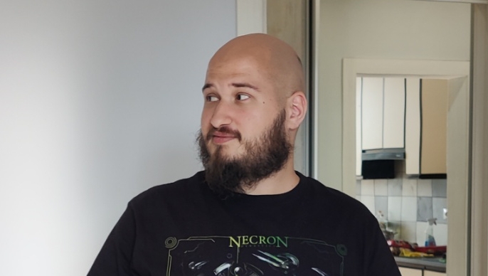 A man with a shaved head and full beard wearing a black "Necron" graphic t-shirt looks off to the side with a slight smile, standing indoors with a kitchen visible in the background. The casual portrait captures him from the chest up in what appears to be a residential setting.