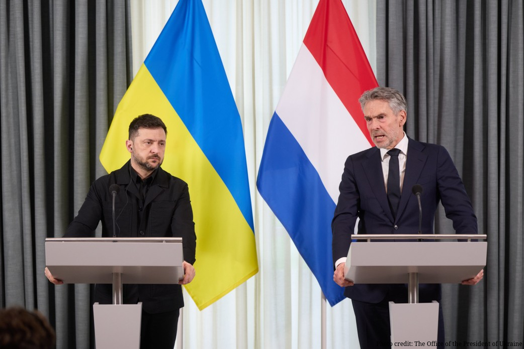 Two leaders stand at separate podiums during a joint press conference, with the Ukrainian flag (blue and yellow) and Dutch flag (red, white and blue) displayed between them against a backdrop of gray and white curtains. The official on the left wears all black, while the official on the right wears a dark suit with white shirt and tie, photo credited to The Office of the President of Ukraine.