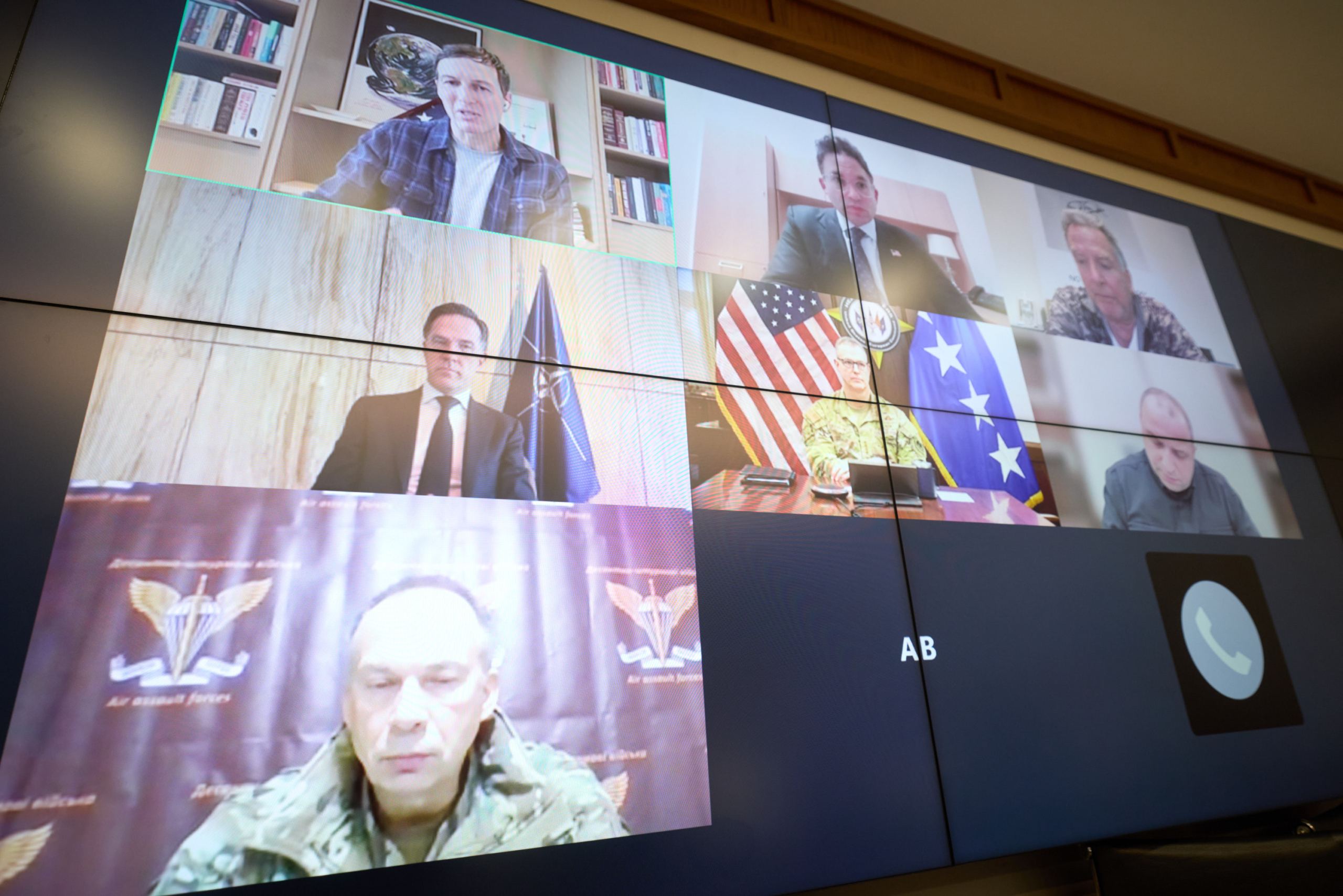 A video conference display shows multiple participants in a grid layout, including government officials with the American flag visible in one frame, and a military officer in camouflage uniform in another. The large screen captures what appears to be an official diplomatic or military meeting conducted virtually.