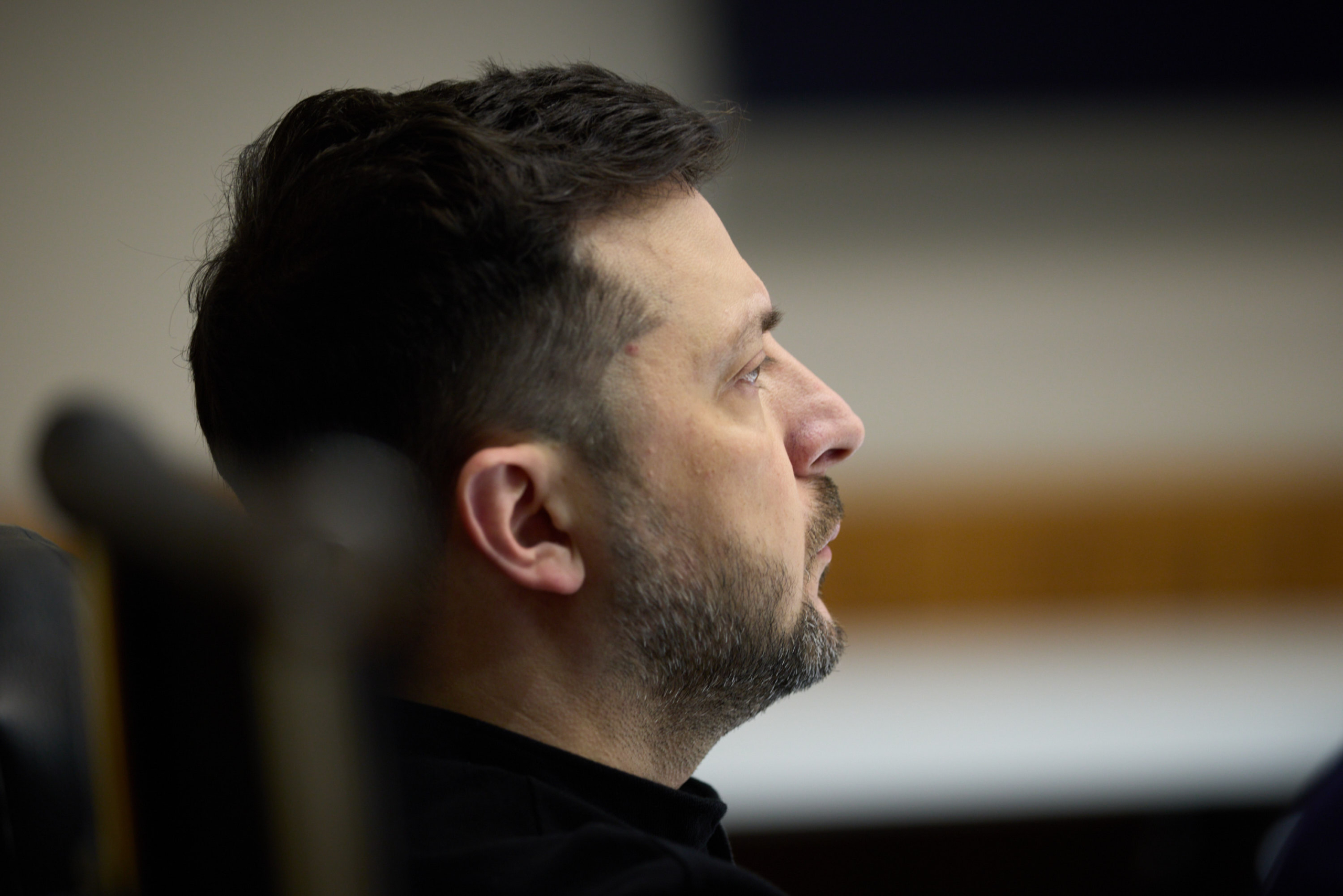 A bearded man in a dark shirt shown in profile, looking upward with a slight smile in what appears to be an indoor setting. The shallow depth of field creates a blurred background with warm tones.