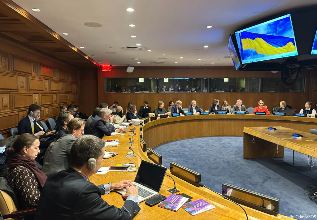 Diplomats and officials sit around a curved wooden conference table in a formal meeting room, with multiple overhead screens displaying the Ukrainian flag in blue and yellow. The wood-paneled chamber features recessed lighting and a viewing gallery visible in the background.