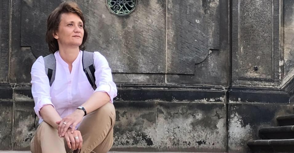 A woman wearing a white shirt, beige pants, and sandals sits on stone steps with a backpack, positioned in front of a weathered wall featuring decorative metalwork. The casual outdoor setting shows worn architectural details including aged metal panels and railings.