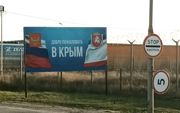 A roadside billboard reading "Welcome to Crimea" in Russian displays the Russian flag and Crimean emblem, positioned near a stop sign marked "CONTROL" and a speed limit sign showing 5, with fencing and dry grassland visible in the background.