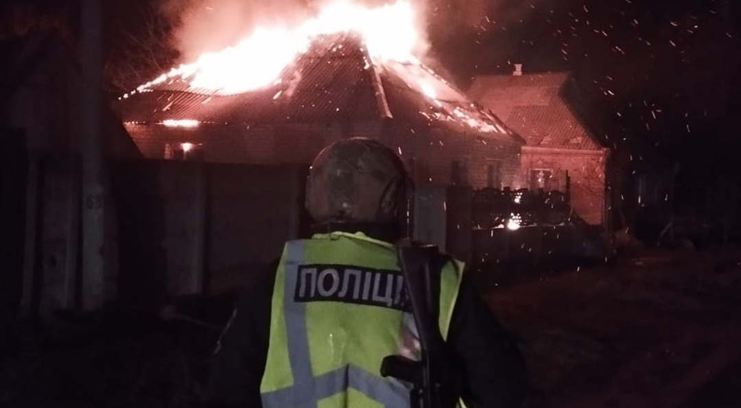 A police officer in a reflective yellow vest with "ПОЛІЦІЯ" (Police) text stands watching a residential building engulfed in intense flames at night, with embers flying through the dark sky. The National Police of Ukraine logo appears in the bottom right corner.