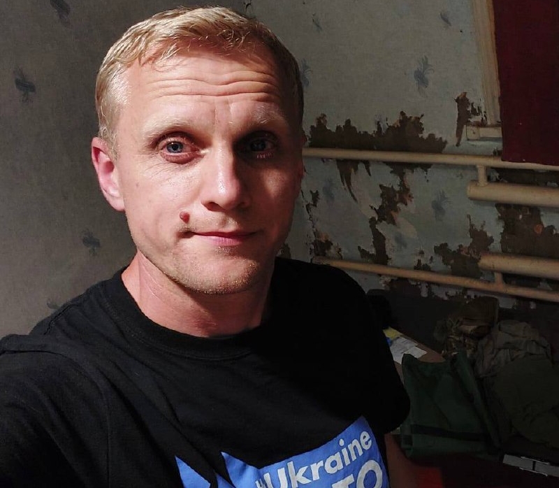 A man with short blonde hair and light stubble wears a black t-shirt with "Ukraine" text in blue and white, taking a selfie indoors against a weathered wall with peeling paint and exposed wooden slats. His direct gaze and slight smile create an informal portrait in what appears to be a damaged or aged interior space.