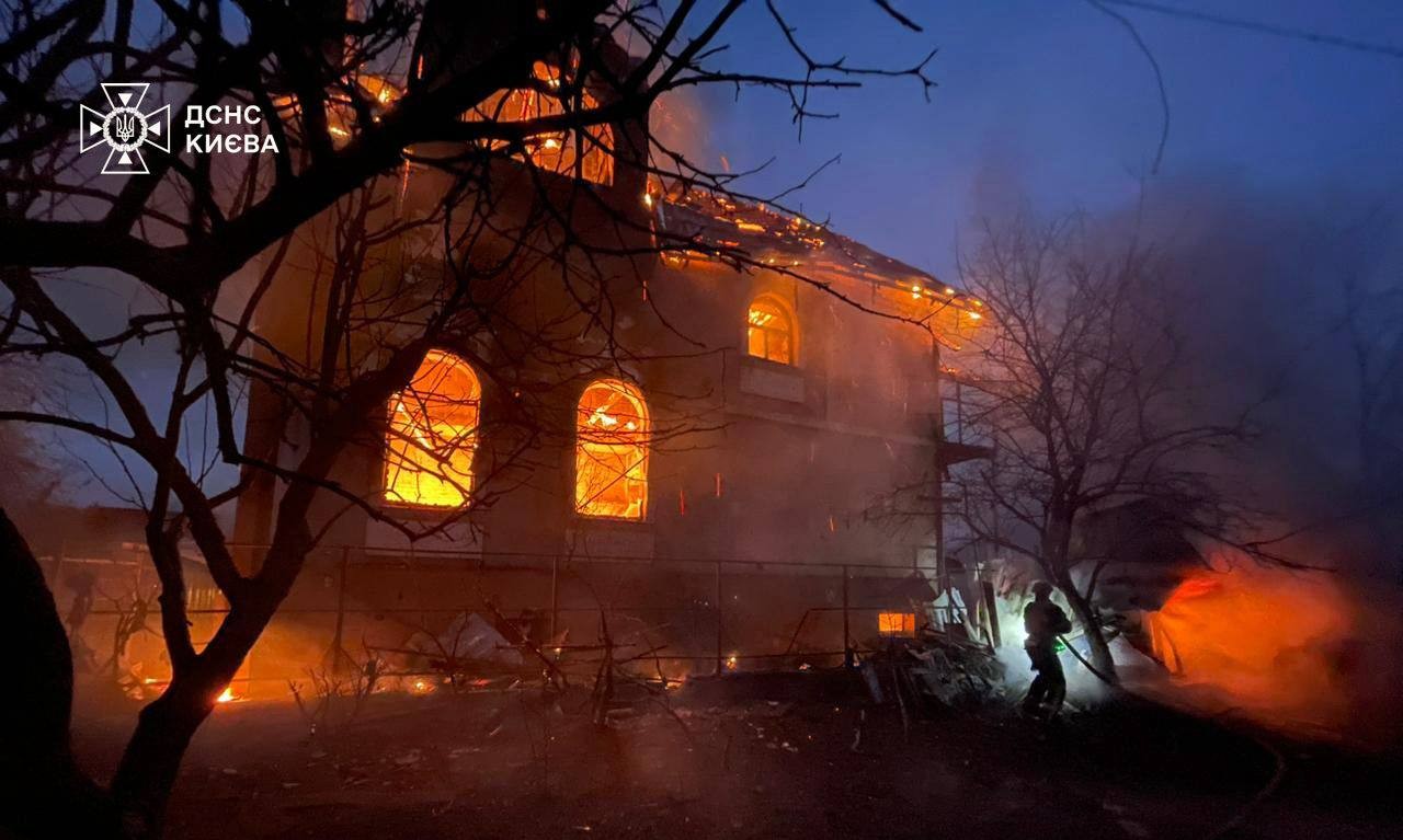Firefighters battle a massive blaze engulfing a historic building at dusk, with flames visible through arched windows and the roof structure as smoke billows into the darkening sky. The State Emergency Service of Kyiv logo appears in the top left corner, documenting the emergency response to the burning structure silhouetted by bare trees.