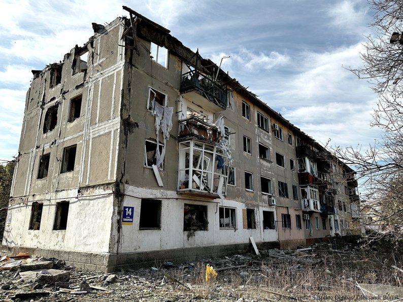 A severely damaged four-story residential building with its roof completely destroyed, exposing charred structural beams, and multiple blown-out windows throughout the facade stands against a cloudy sky. The war-torn apartment block shows extensive blast damage, with debris scattered at its base and bare trees visible nearby, while the ground floor displays a blue street number sign amid the devastation.