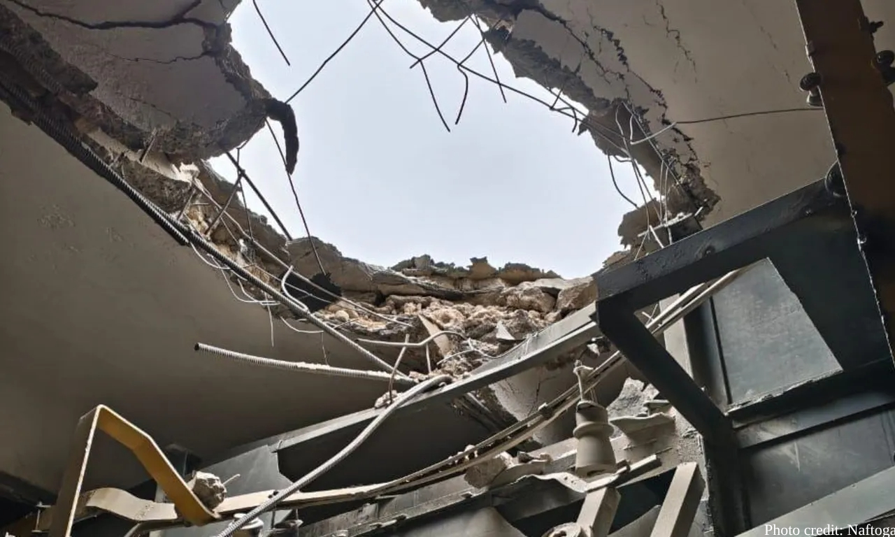 A massive hole in a building's ceiling reveals the sky above, with twisted rebar, broken concrete, collapsed structural elements and dangling wires visible amid the destruction. The view looks upward through multiple floors that have been blown through, with rubble and damaged infrastructure filling the frame, photo credited to Naftogaz.