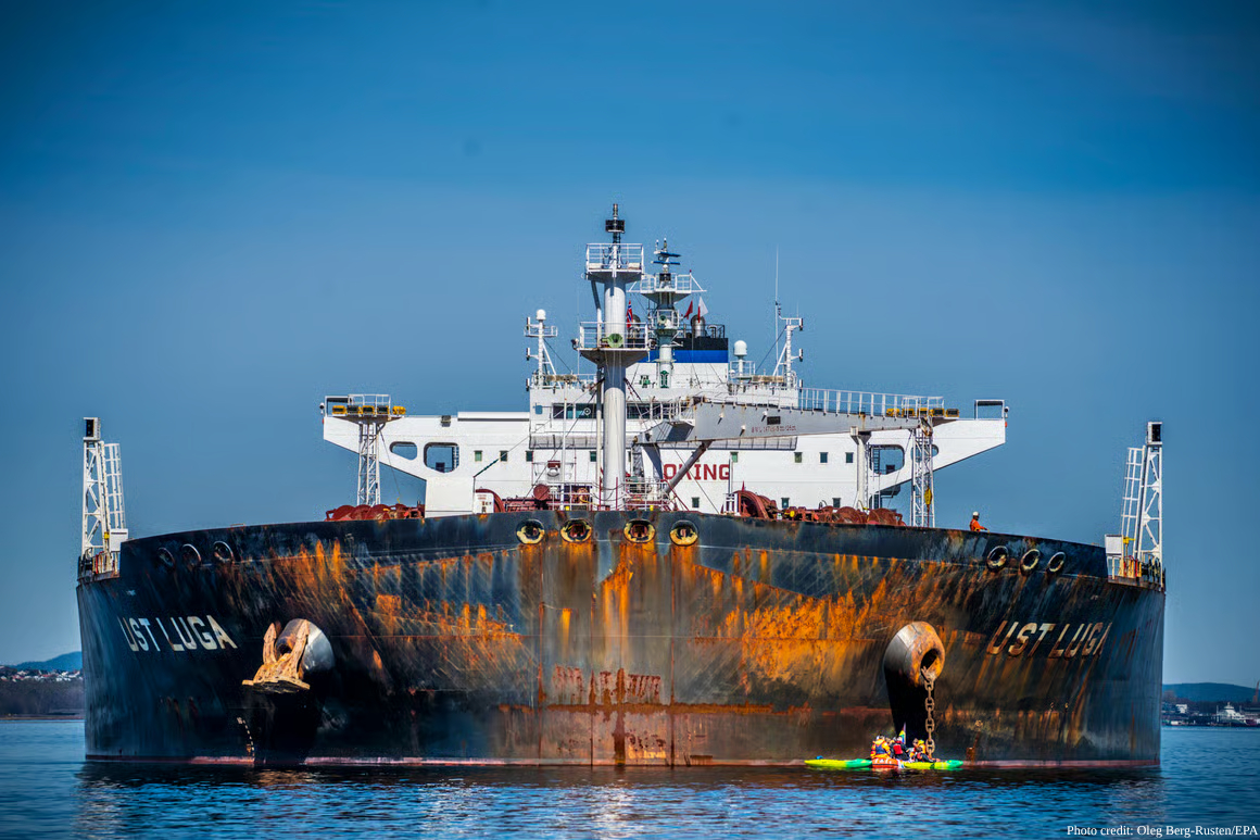 A heavily rusted tanker vessel named "HST LUGA" sits at sea with visible orange-brown corrosion streaking down its black hull, while kayakers paddle in yellow boats near its bow. The ship's white superstructure contrasts with its weathered dark hull beneath clear blue skies, photo credited to Oleg Berg-Rusten/EPA.
