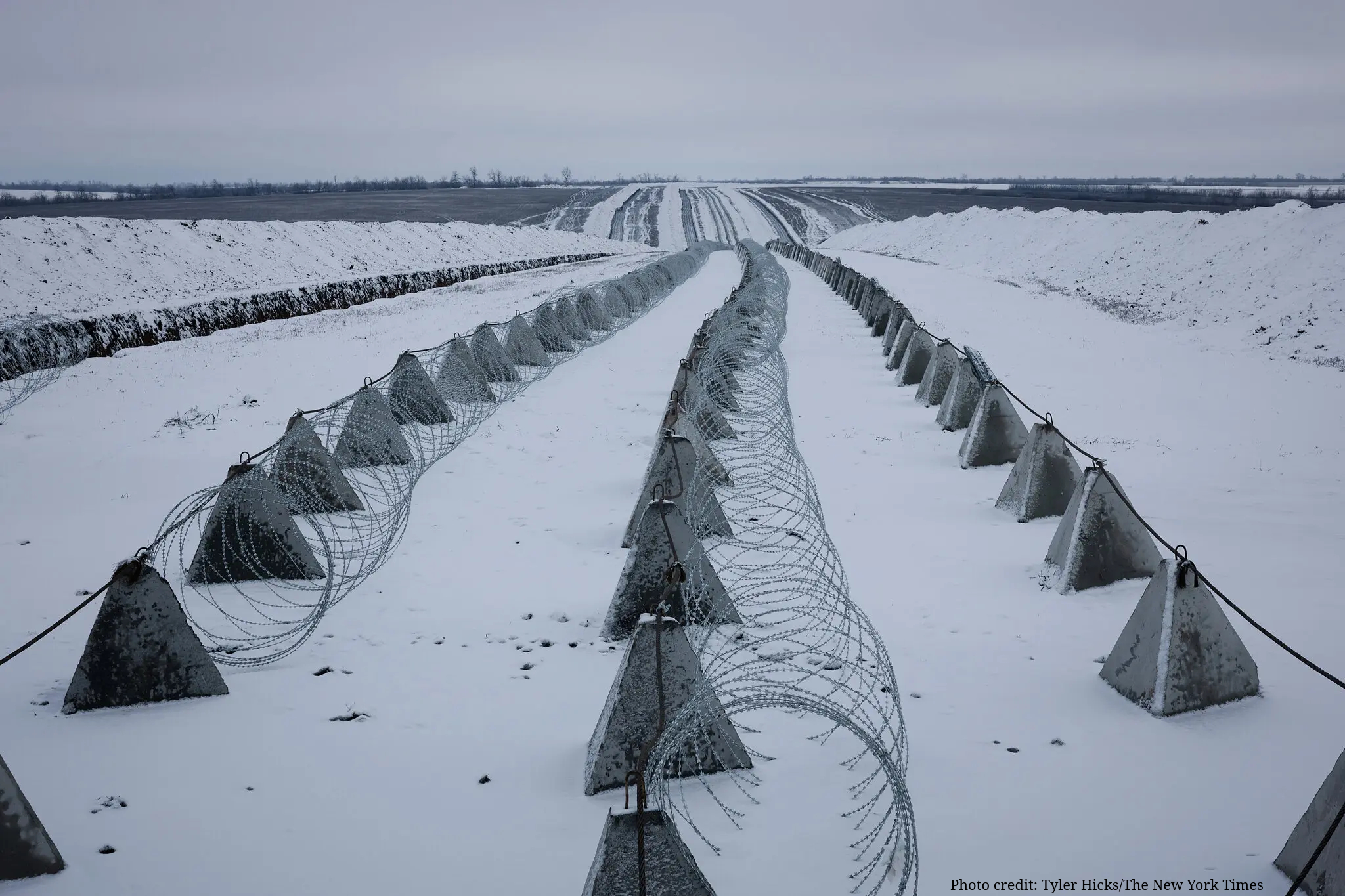 Three long, parallel rows of concrete dragon’s teeth anti-tank obstacles and coils of razor wire stretch across a snow-covered field under a grey sky. The fortifications disappear into the distance toward a flat horizon.