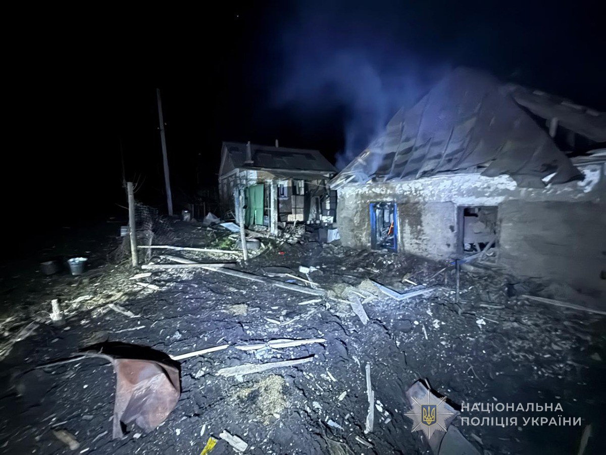 Two severely damaged buildings stand at night with collapsed roofs and walls, debris scattered across the ground, smoke or dust visible in the illuminated air, and a blue doorway visible in the structure on the right, documented by the National Police of Ukraine.
