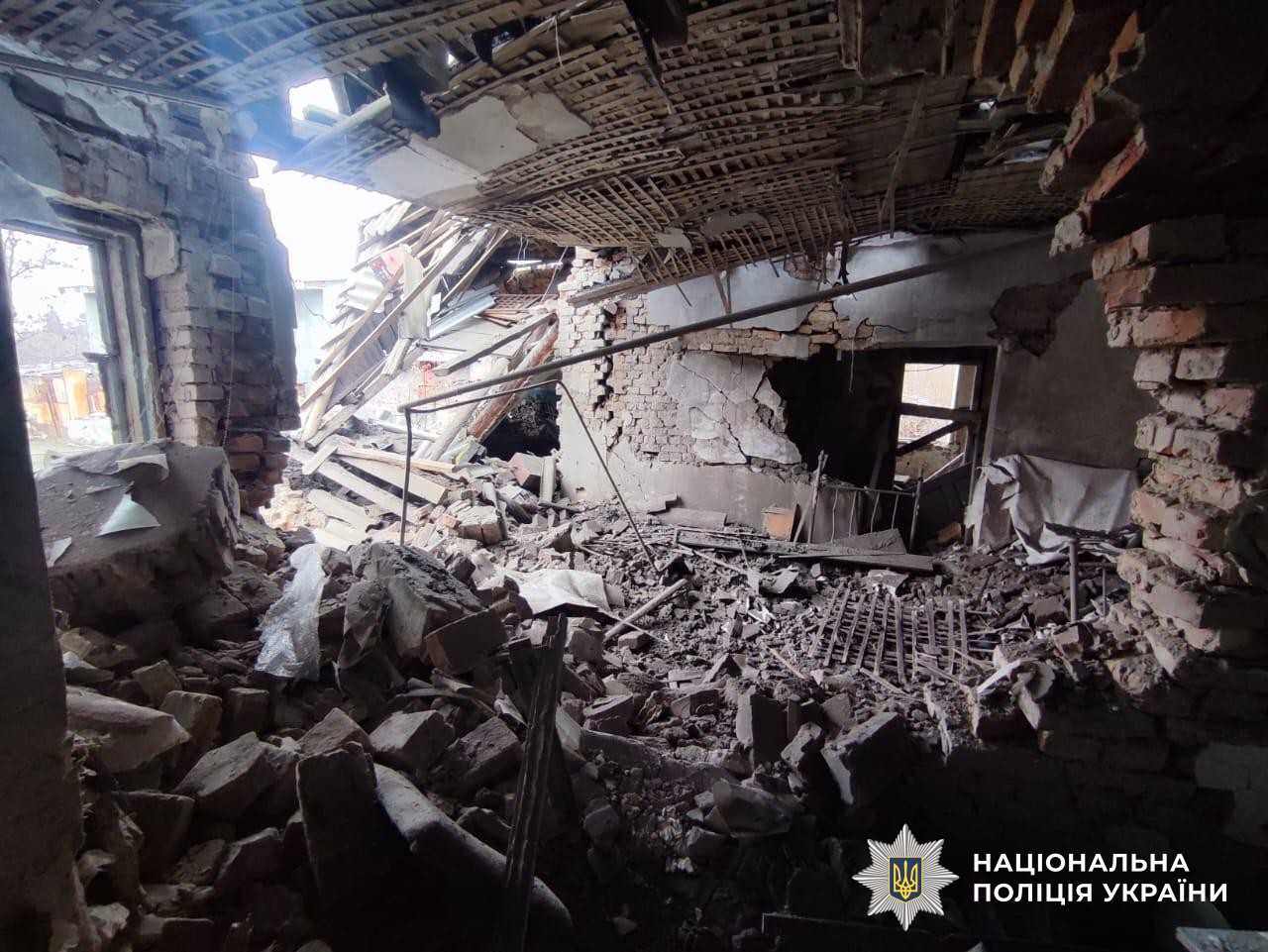 Interior of a severely damaged building shows collapsed ceiling with exposed wooden lattice work, destroyed brick walls, piles of rubble covering the floor, broken metal bed frame, and sunlight streaming through large openings where walls once stood, documented by the National Police of Ukraine.