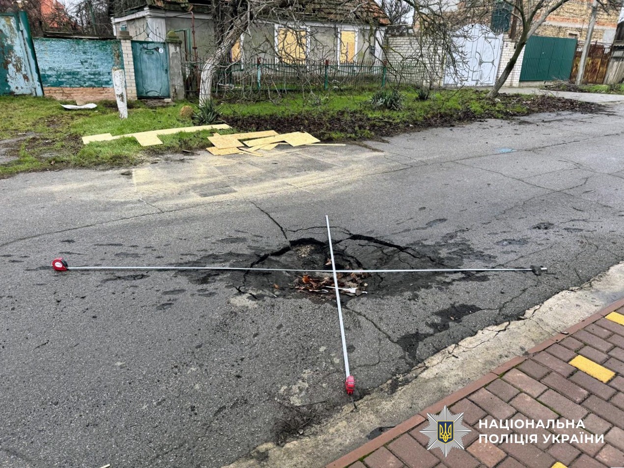 A blast crater in an asphalt street is marked with white measurement markers forming a cross pattern, with scattered yellow insulation panels on grass, residential buildings with bare trees in the background, and National Police of Ukraine logo in the corner.