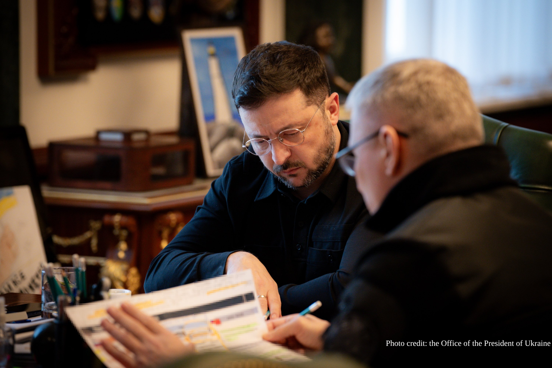 Two men in an office setting review documents together at a desk, with the man in the foreground wearing glasses and a dark navy shirt holding papers while the other person in black looks on, surrounded by office furniture and a framed blue artwork in the background.