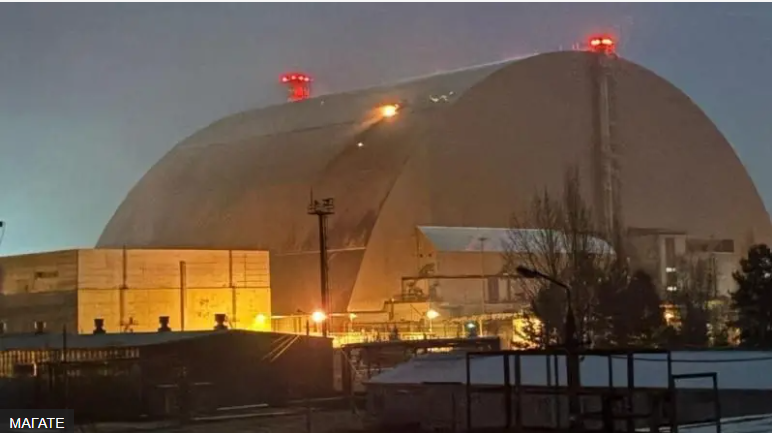 The massive New Safe Confinement structure dominates the Chernobyl Nuclear Power Plant at dusk, its curved metallic dome illuminated by lights and topped with red warning beacons. The industrial facility features yellow-lit buildings in the foreground and bare winter trees silhouetted against the evening sky.