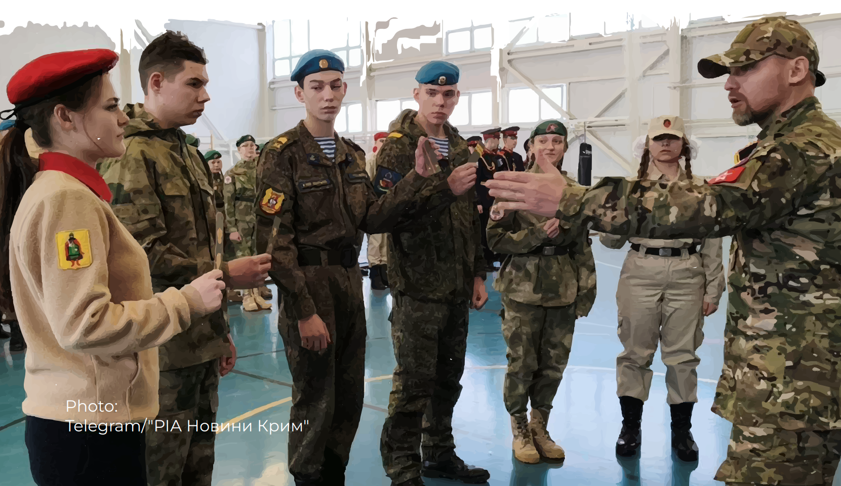 A military instructor in camouflage and a tan cap demonstrates something to a group of Ukrainian children wearing various military uniforms and berets inside a occupied by Russia Ukrainian school. The cadets, including both males and a female in a tan uniform with a red beret, stand at attention while receiving instruction.