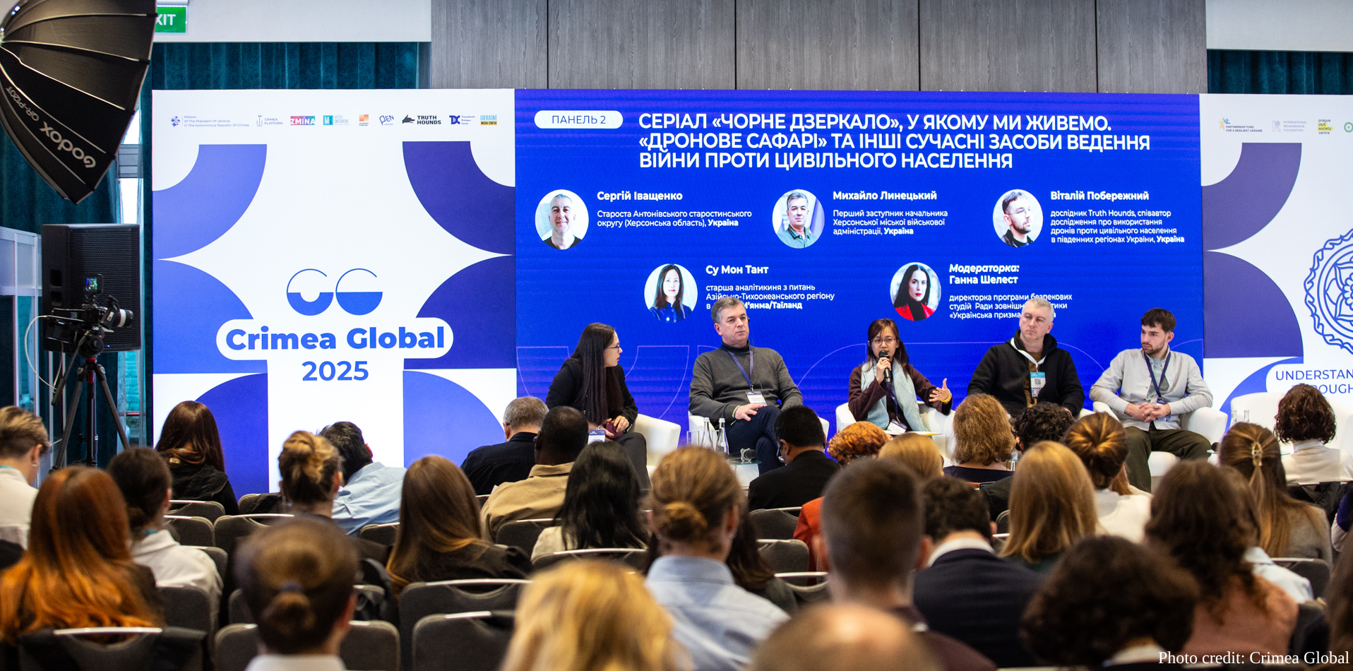 A panel discussion takes place on stage at Crimea Global 2025, with five speakers seated in white chairs before an audience. The vibrant blue backdrop displays participant profiles and Cyrillic text about a discussion panel concerning civilian population protection during wartime.