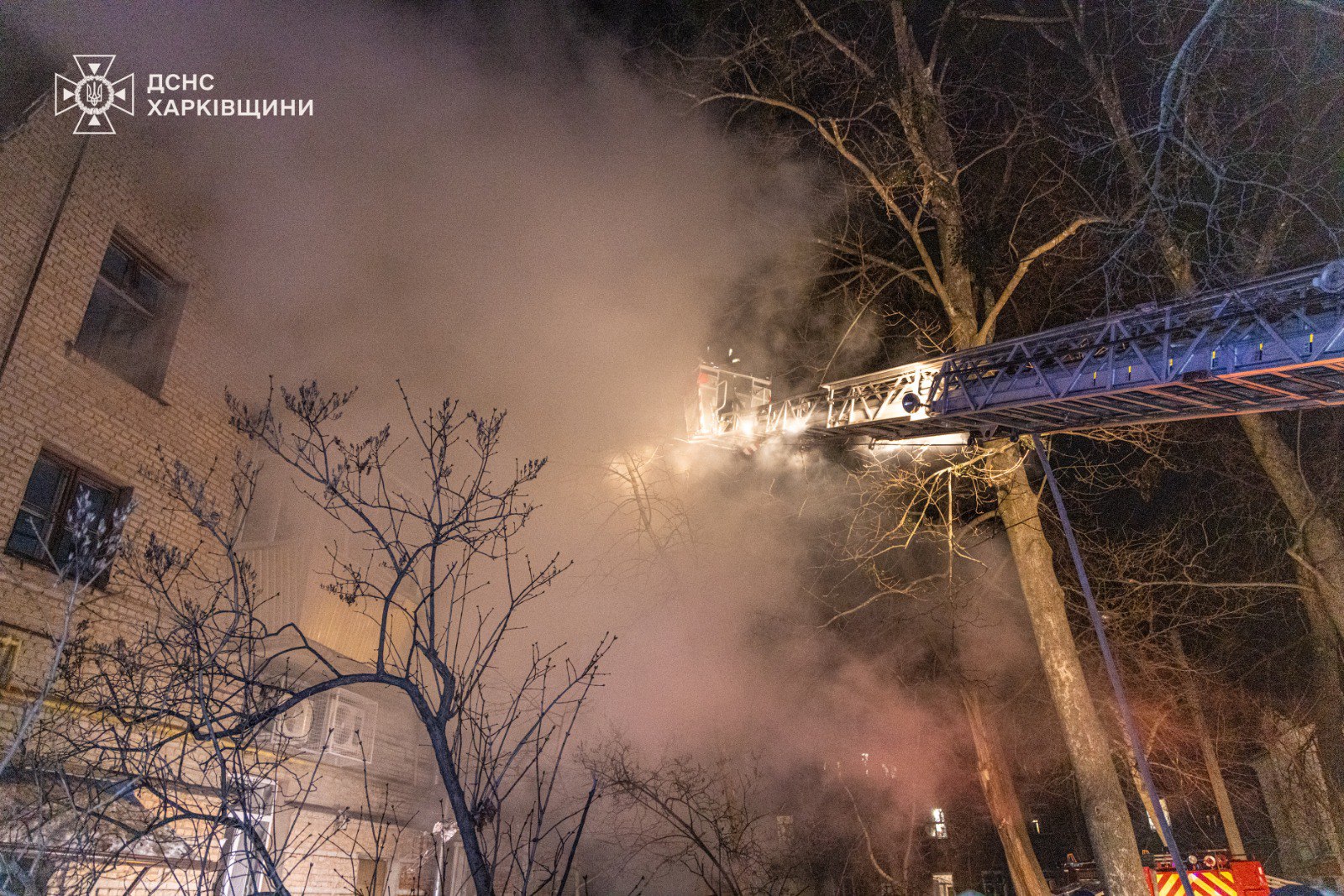 Firefighters battle a nighttime blaze from an extended aerial ladder positioned near bare winter trees, with thick smoke billowing past a brick apartment building. Emergency crews illuminate the scene while working to extinguish the fire in challenging conditions.