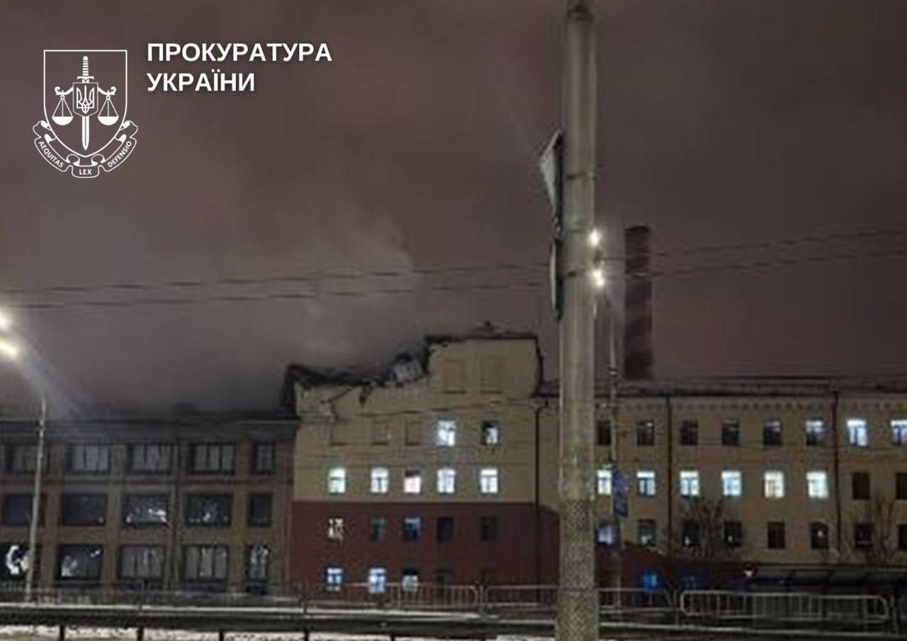 Multi-story residential buildings of Roshen factory in Kyiv at dusk with illuminated windows show significant structural damage, particularly visible dark damage to the roof and upper sections of the central beige building, with the Prosecutor's Office of Ukraine logo and text overlaid in the upper left corner. Street lights and barriers are visible in the foreground along with bare winter trees against an overcast evening sky.