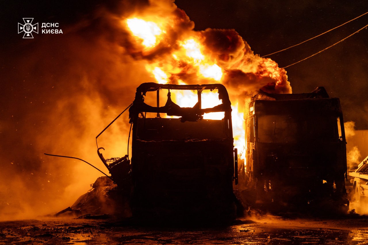 A vehicle burns intensely at night, completely engulfed in towering orange flames and thick black smoke. The skeletal frame of the vehicle is silhouetted against the brilliant blaze, with its windows blown out and interior completely consumed by fire.