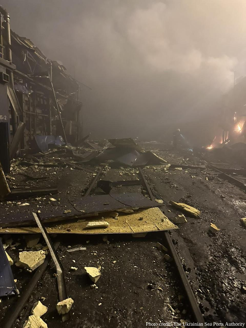 A wide area of industrial or port infrastructure is covered in scattered debris, twisted metal, and insulation panels at night under thick smoke. A lone emergency worker is visible in the distance with a flashlight, and a photo credit for The Ukrainian Sea Ports Authority is in the bottom right.