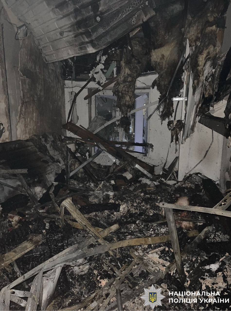 The interior of a room is heavily damaged and charred by fire, with debris, broken wooden beams, and insulation hanging from the ceiling. A logo for the National Police of Ukraine is visible in the bottom right corner against the wreckage.