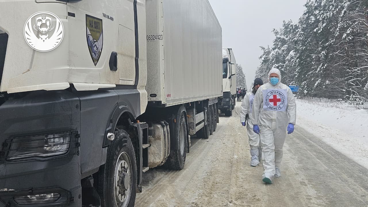 A line of white International Committee of the Red Cross (ICRC) semi-trucks travels down a snow-covered road lined with frosted pine trees. Two personnel in white protective suits and face masks walk along the road beside the lead vehicle, which displays an ICRC emblem and humanitarian markings.