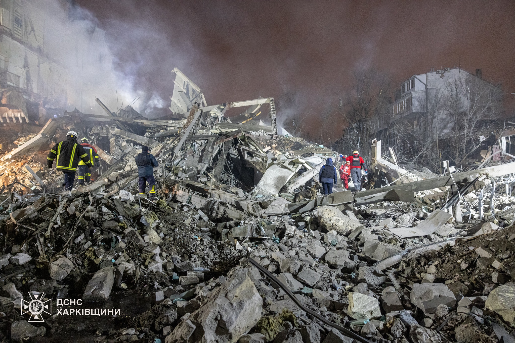 Emergency responders and rescue workers navigate through massive piles of concrete rubble and twisted debris from a completely destroyed building, with thick smoke filling the air and damaged structures visible in the background. Firefighters in reflective gear and rescue personnel in red vests work among the devastation as night falls on the catastrophic scene.