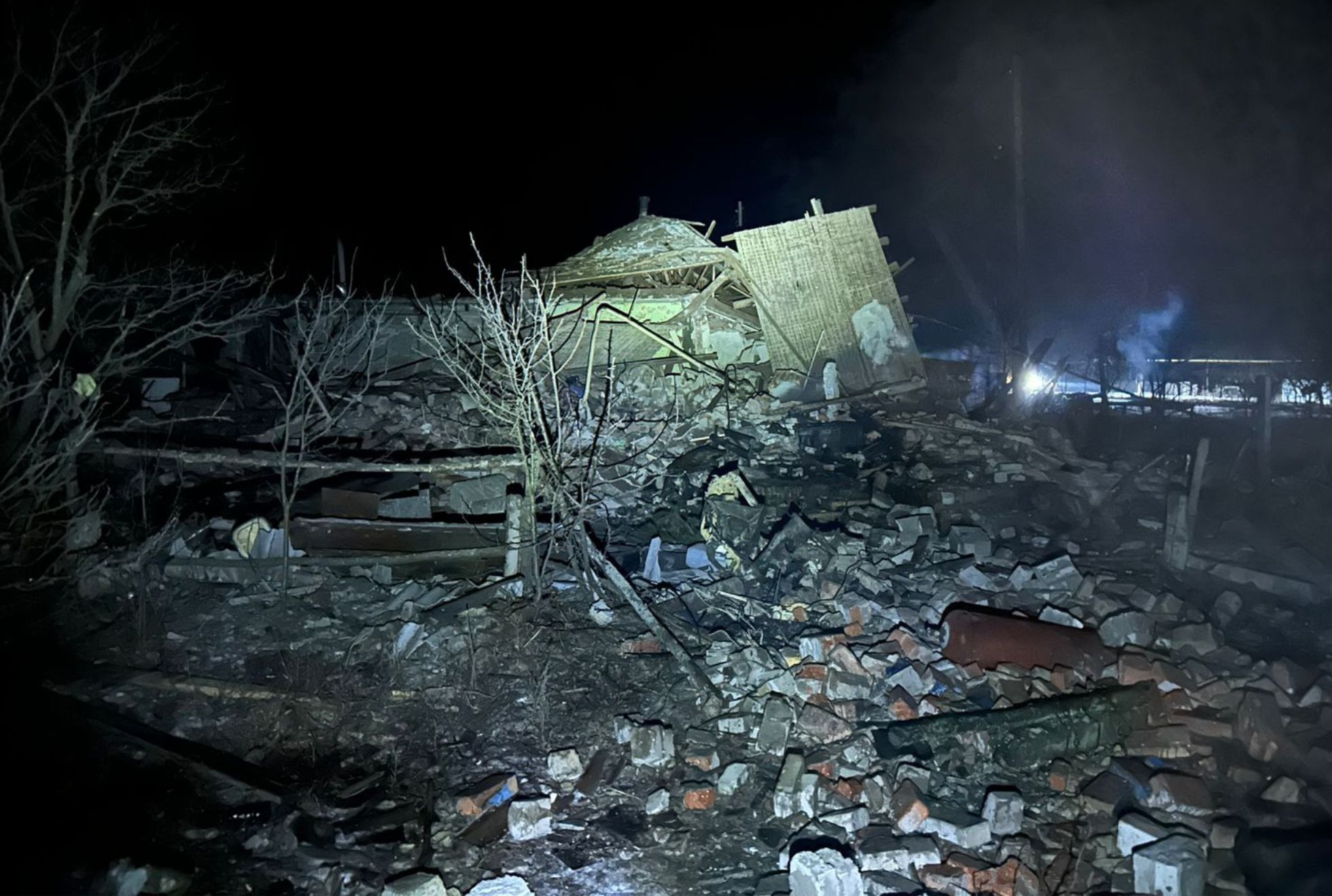Debris and broken bricks cover the ground around a partially collapsed building at night, with damaged walls, roof fragments, and twisted metal visible under artificial light. Bare trees stand amid the rubble, and a faint glow illuminates the background against a night sky.