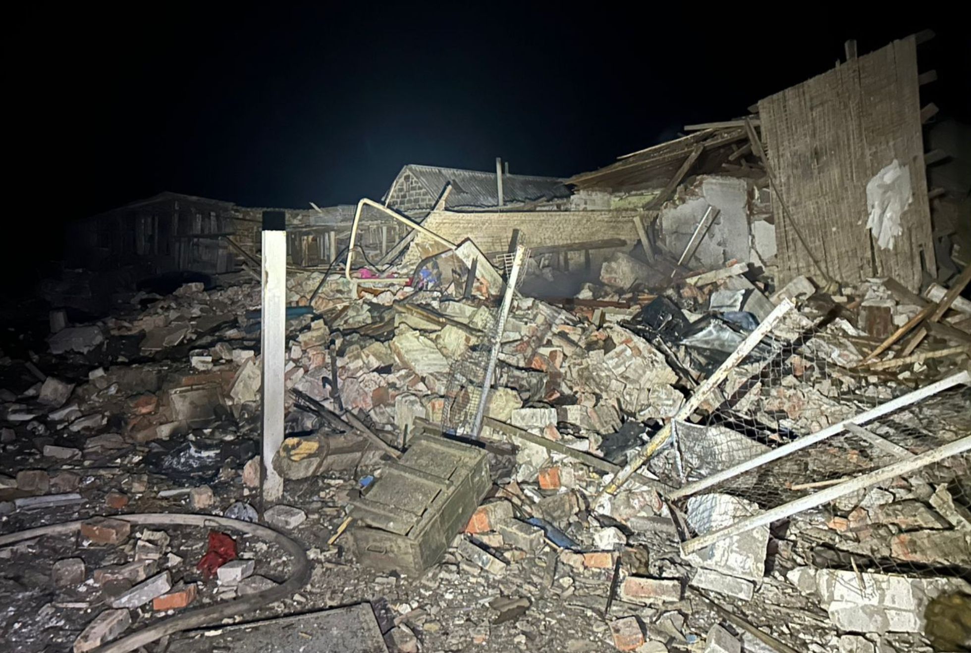 Rubble and debris from a heavily damaged brick building lie scattered across the ground at night, with collapsed walls, broken fencing, and twisted metal visible under artificial light. Partially standing structures and roof fragments remain in the background against a night sky.