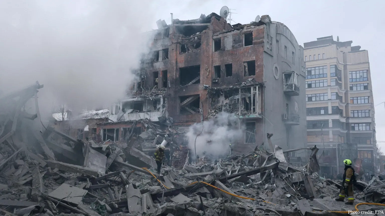 Firefighters in reflective gear work amid massive piles of rubble and debris from a heavily damaged multi-story residential building in Kharkiv with collapsed sections, exposed walls, and blown-out windows as smoke continues to rise from the destruction. Emergency responders with fire hoses are visible in the foreground navigating through concrete fragments and twisted metal, with an adjacent white apartment building showing visible damage in the background.