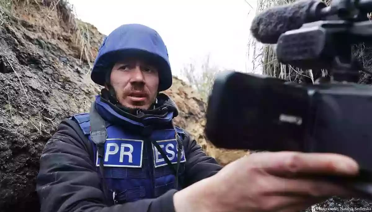 Radio Liberty correspondent Marian Kushnir, wearing a blue helmet and a "PRESS" tactical vest, operates a video camera while positioned in a dirt trench. His face shows signs of minor injury or dirt, and the background consists of the earthen walls of the trench.
