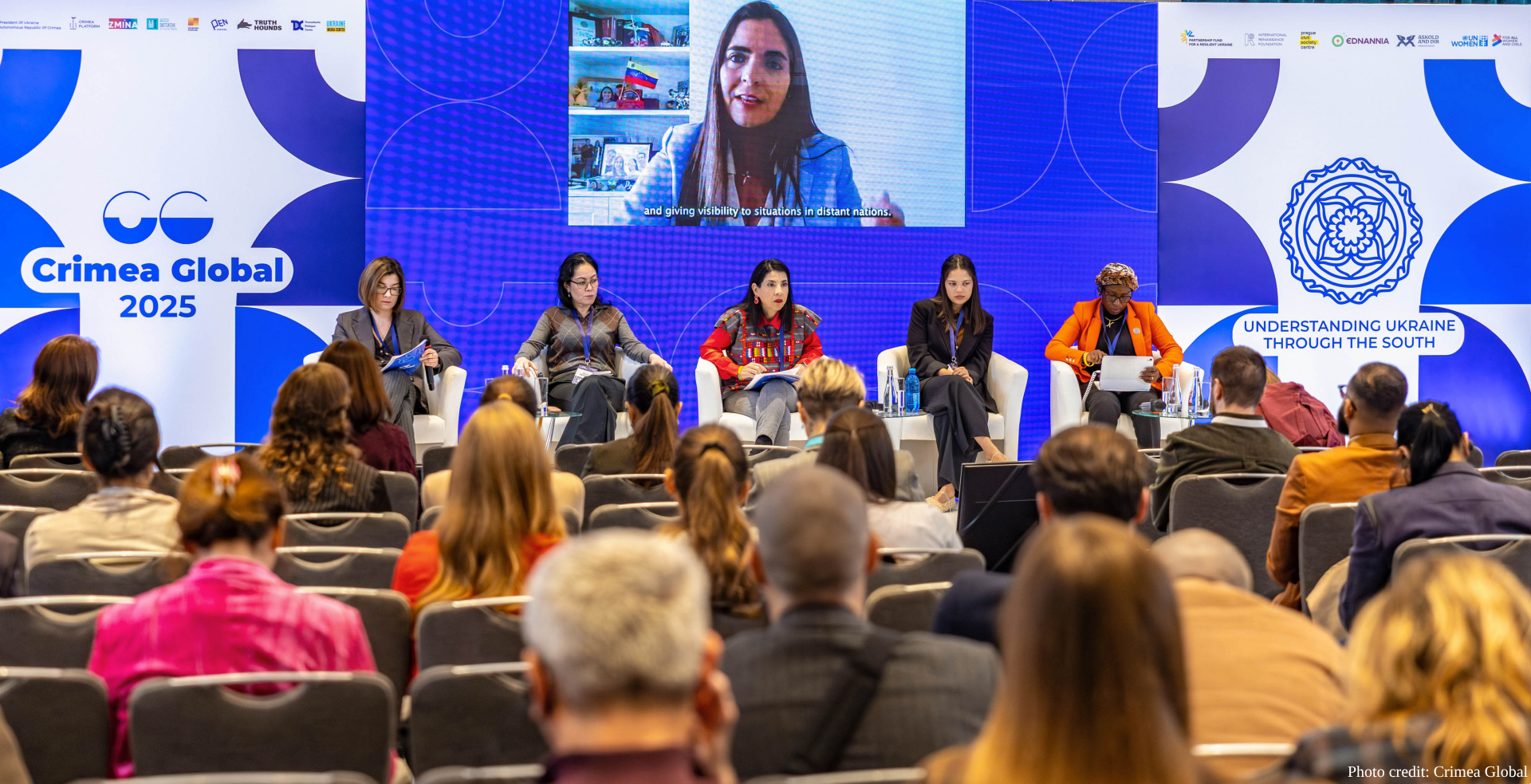 Five female panelists sit in white chairs on a conference stage beneath a large screen displaying a remote participant, with "Crimea Global 2025" and "Understanding Ukraine Through the South" branding visible on blue backdrop panels as an audience watches from rows of chairs. Photo credited to Crimea Global shows the international forum focused on Ukraine-related discussion with diverse speakers and attendees.