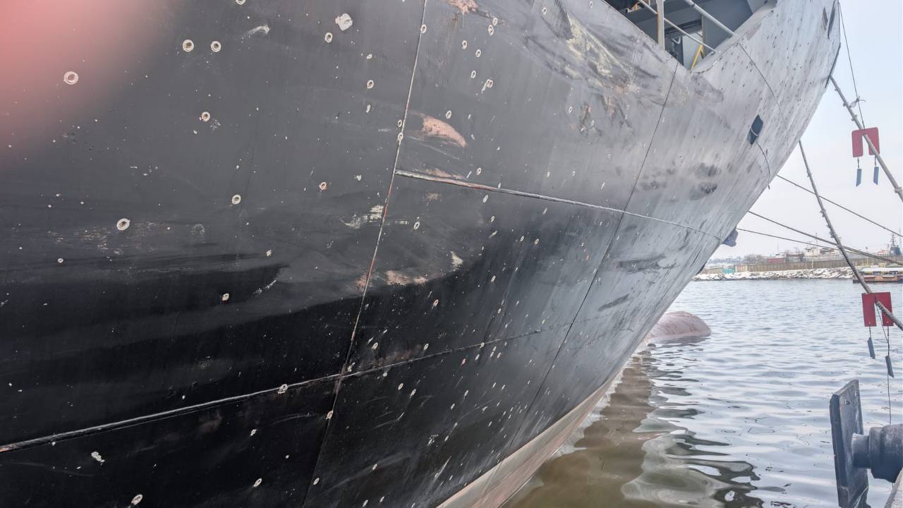 A large dark-hulled naval or cargo vessel displays extensive damage with numerous visible impact marks across its metal surface, moored at a port with calm waters and flags visible on the deck. The ship's weathered exterior shows significant deterioration with rust stains and multiple circular penetration marks scattered across its hull plating.