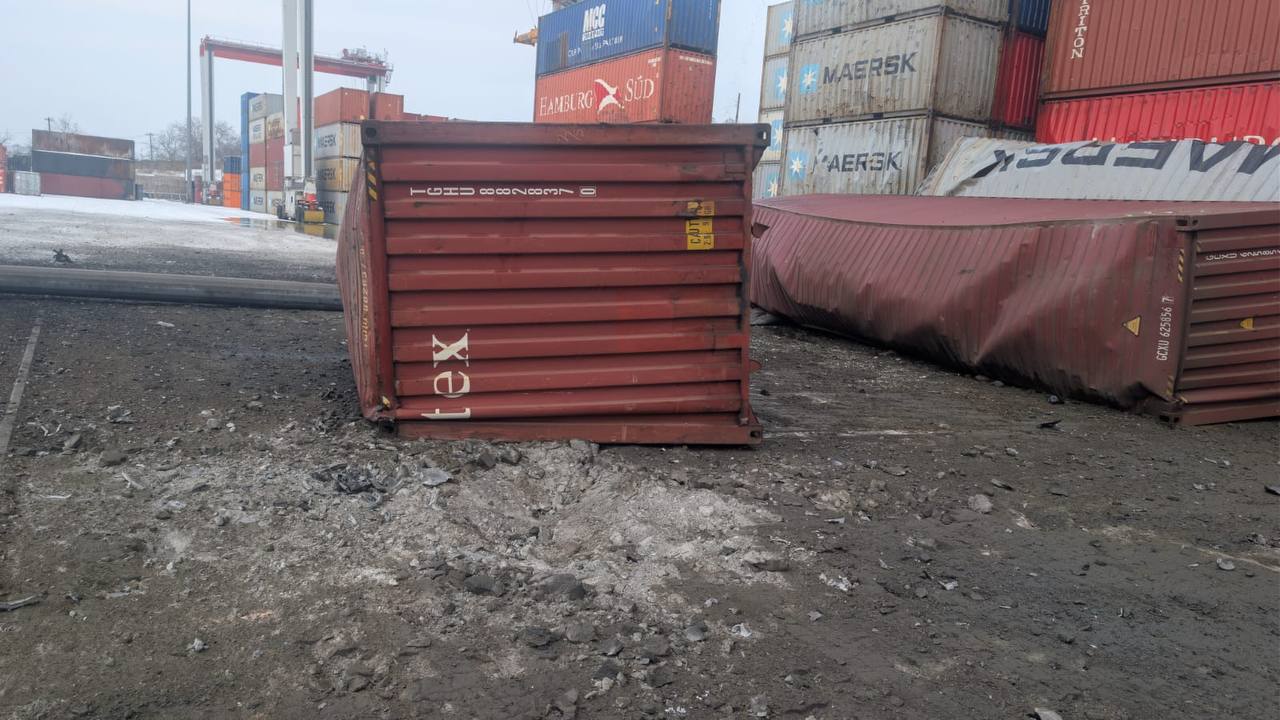A red shipping container lies severely damaged on rough terrain at a container yard, with its side crushed inward. Behind it, stacks of multicolored containers from various shipping companies including Maersk and Hamburg Süd create a backdrop of international cargo infrastructure.
