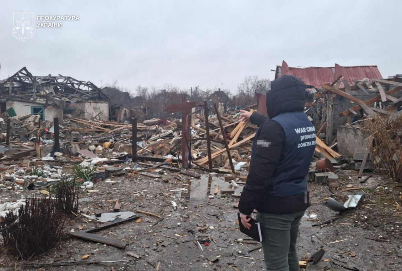 A prosecutor from the Donetsk Regional Prosecutor's Office points toward a site of total destruction where houses have been reduced to piles of wooden beams and rubble. The official, wearing a dark blue vest labeled "WAR CRIMES prosecutor DONETSK REGION," documents the aftermath of the massive shelling on February 12, 2026