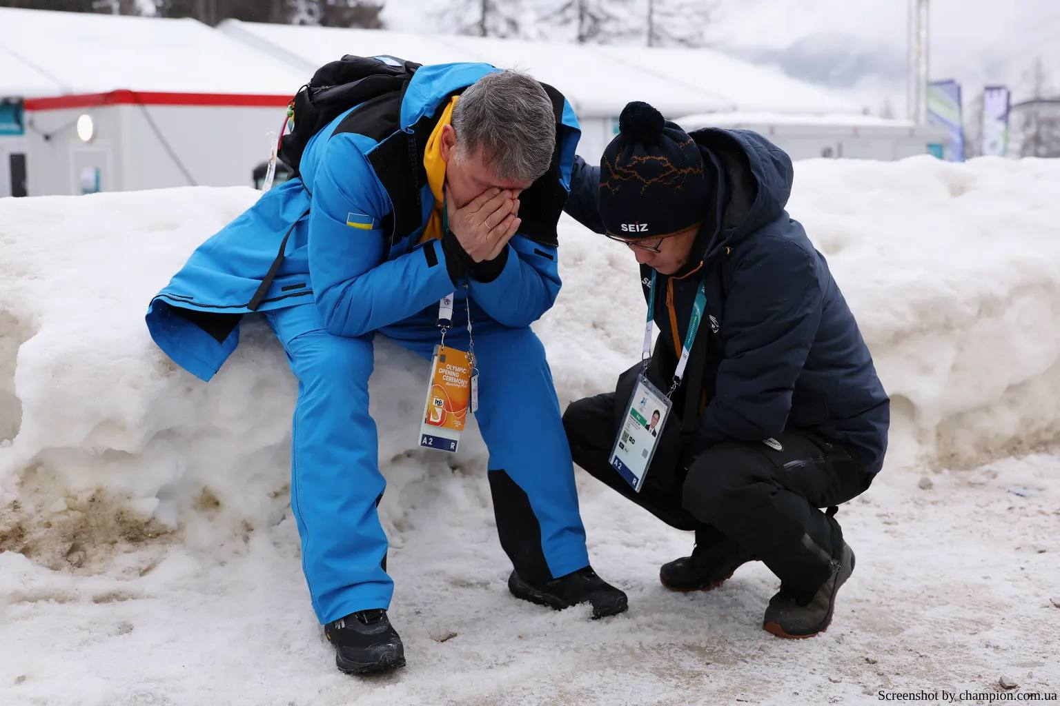 A man wearing a blue and yellow Ukrainian Olympic uniform sits on a snowbank with his head in his hands while another man in a dark winter jacket crouches beside him in support. Both men wear official event credentials around their necks against a backdrop of snow and temporary structures.