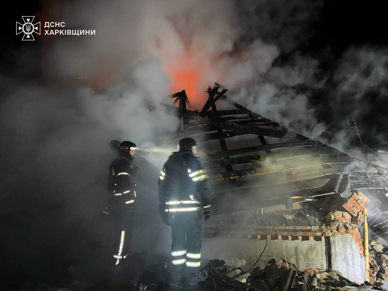 A close-up nighttime shot of a large pile of wooden debris and structural remains burning with bright yellow and orange flames. Thick smoke rises into the black sky. The logo for the State Emergency Service of Ukraine (DSNS) in the Kharkiv region is in the top-left corner.