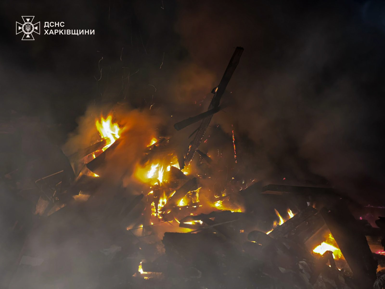 A close-up nighttime shot of a large pile of wooden debris and structural remains burning with bright yellow flames. Thick smoke rises into the black sky. The logo for the State Emergency Service of Ukraine (DSNS) of the Kharkiv region is in the top-left.