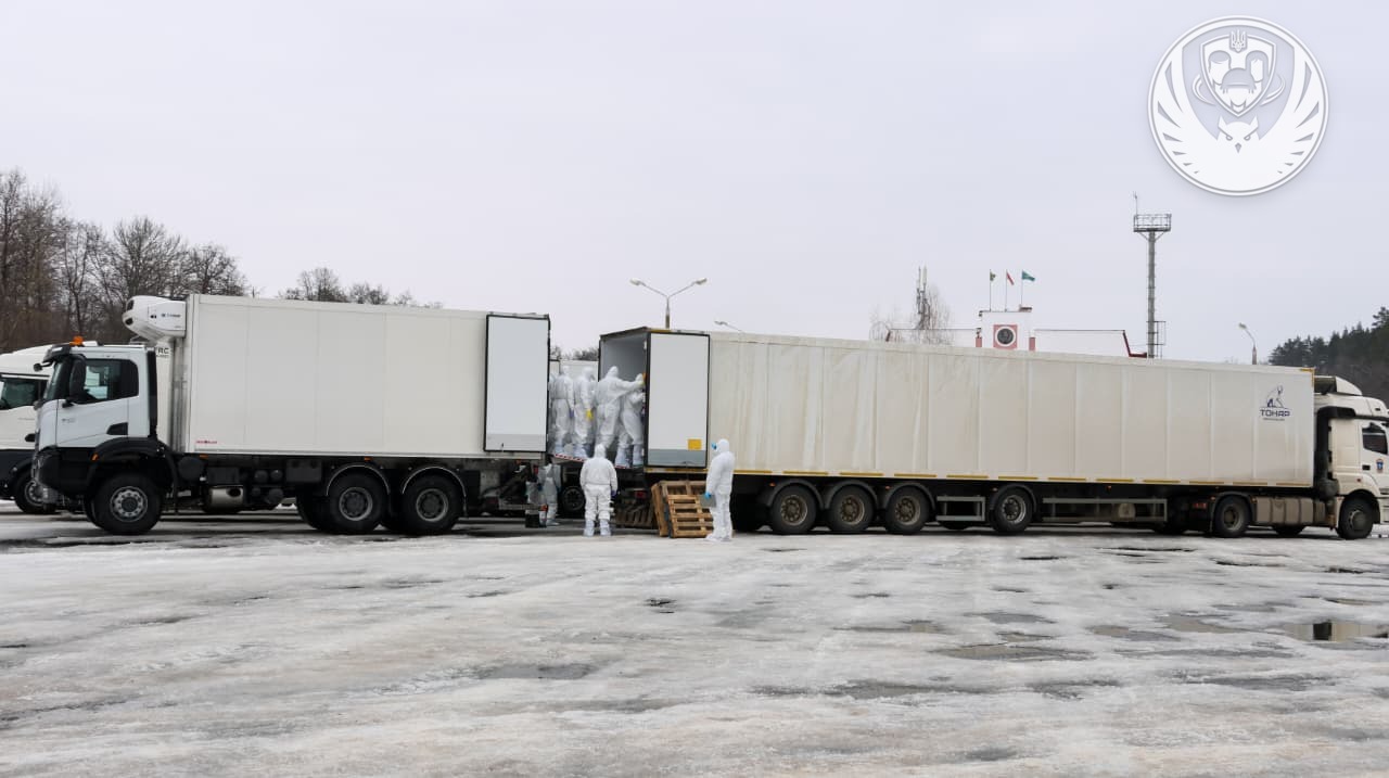 Under a grey winter sky, personnel in white full-body hazmat suits transfer remains between two large white refrigerated trucks in an open, snow-dusted lot. The emblem of the Coordination Headquarters for the Treatment of Prisoners of War is visible in the top-right corner, documenting a repatriation mission in February 2026.